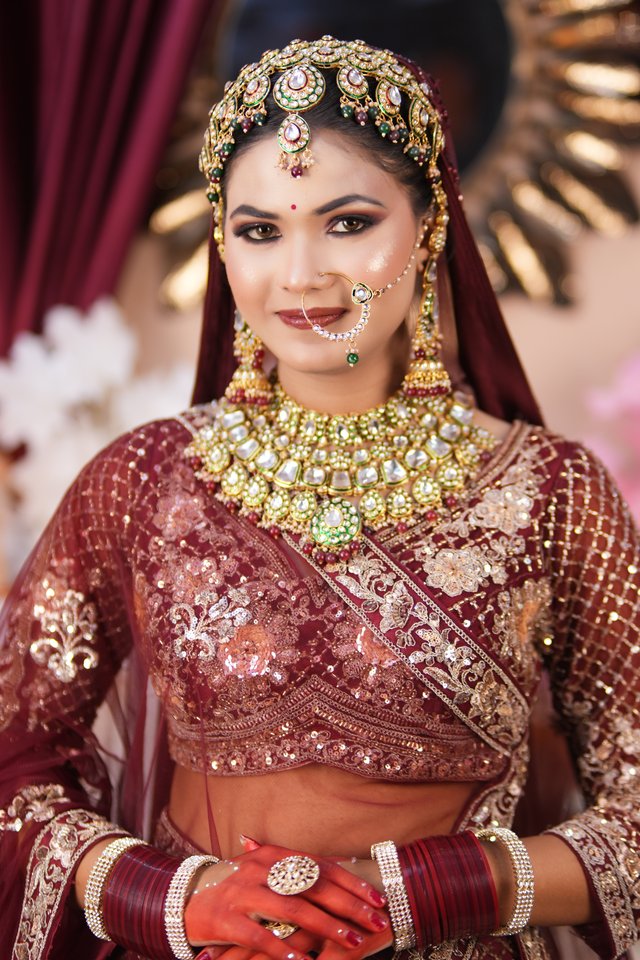 Bride in a maroon bridal lehenga with kundan and pearl jewellery, facing camera with a soft smile